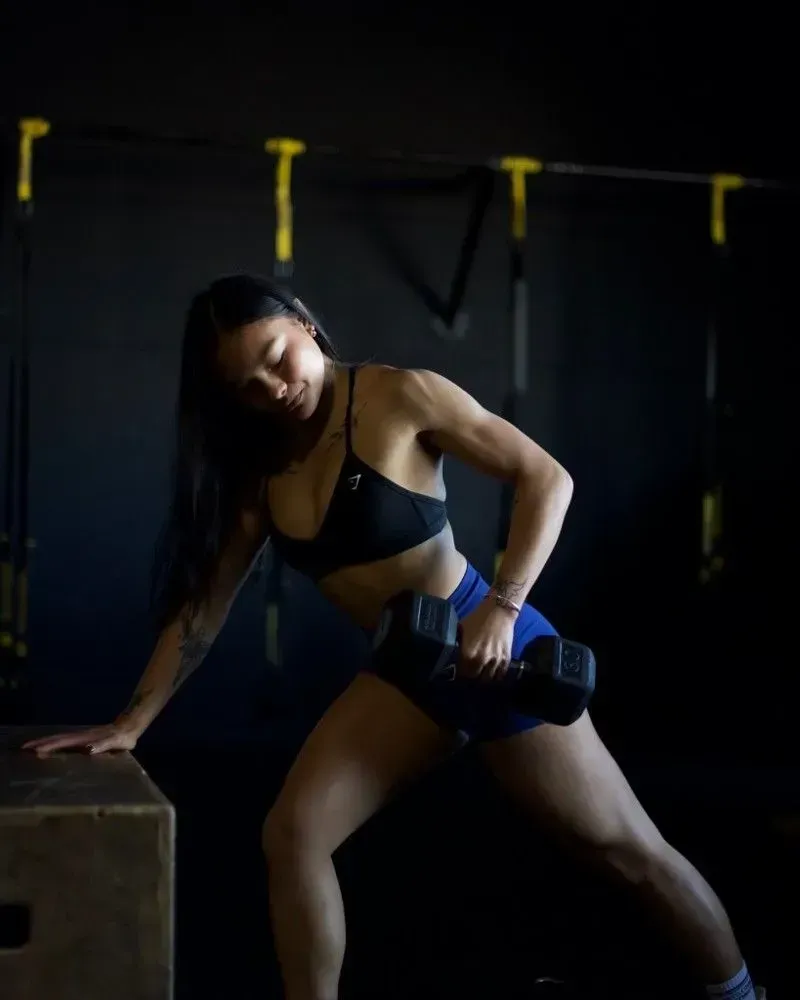 Woman in gym lifting dumbbell, leaning on a box, wearing athletic clothing.