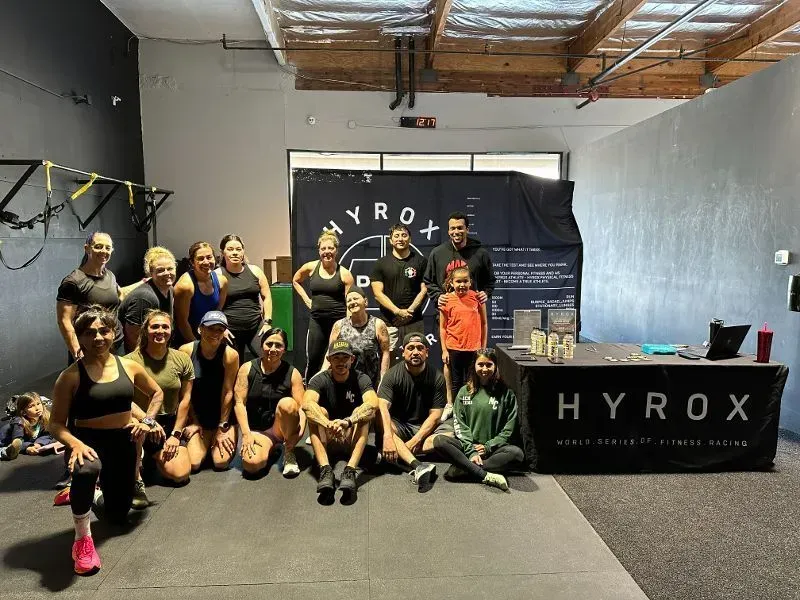 Group of people posing with HYROX banner in a gym setting. Some are kneeling, smiling.