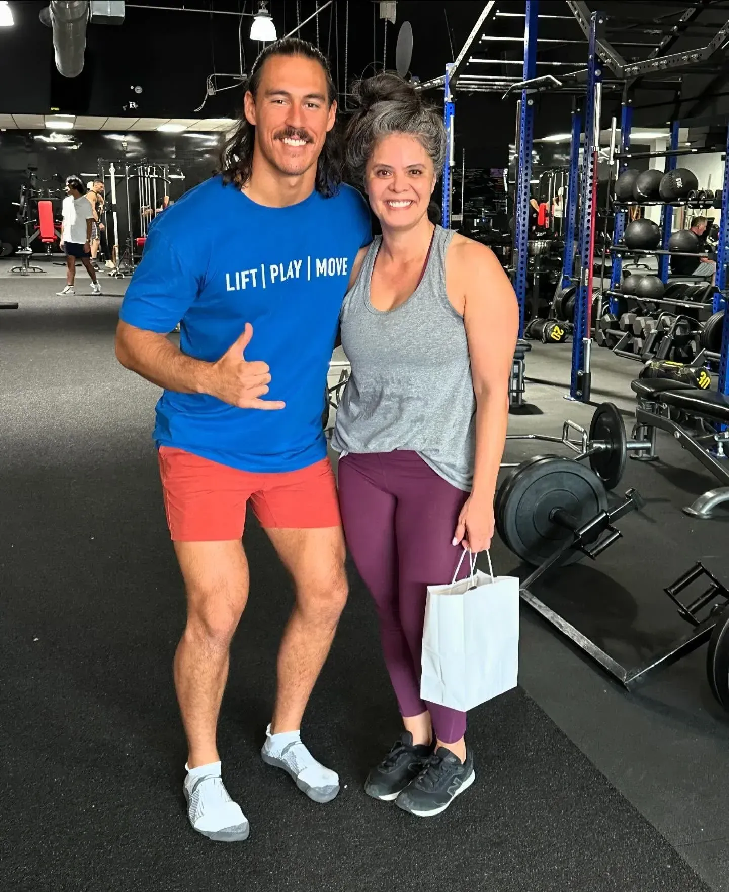 Man in blue shirt gives thumbs up next to a woman in a gym. Both smile.