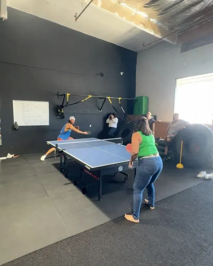 Two people playing ping pong in a gym, others watching, black and gray walls, blue table.