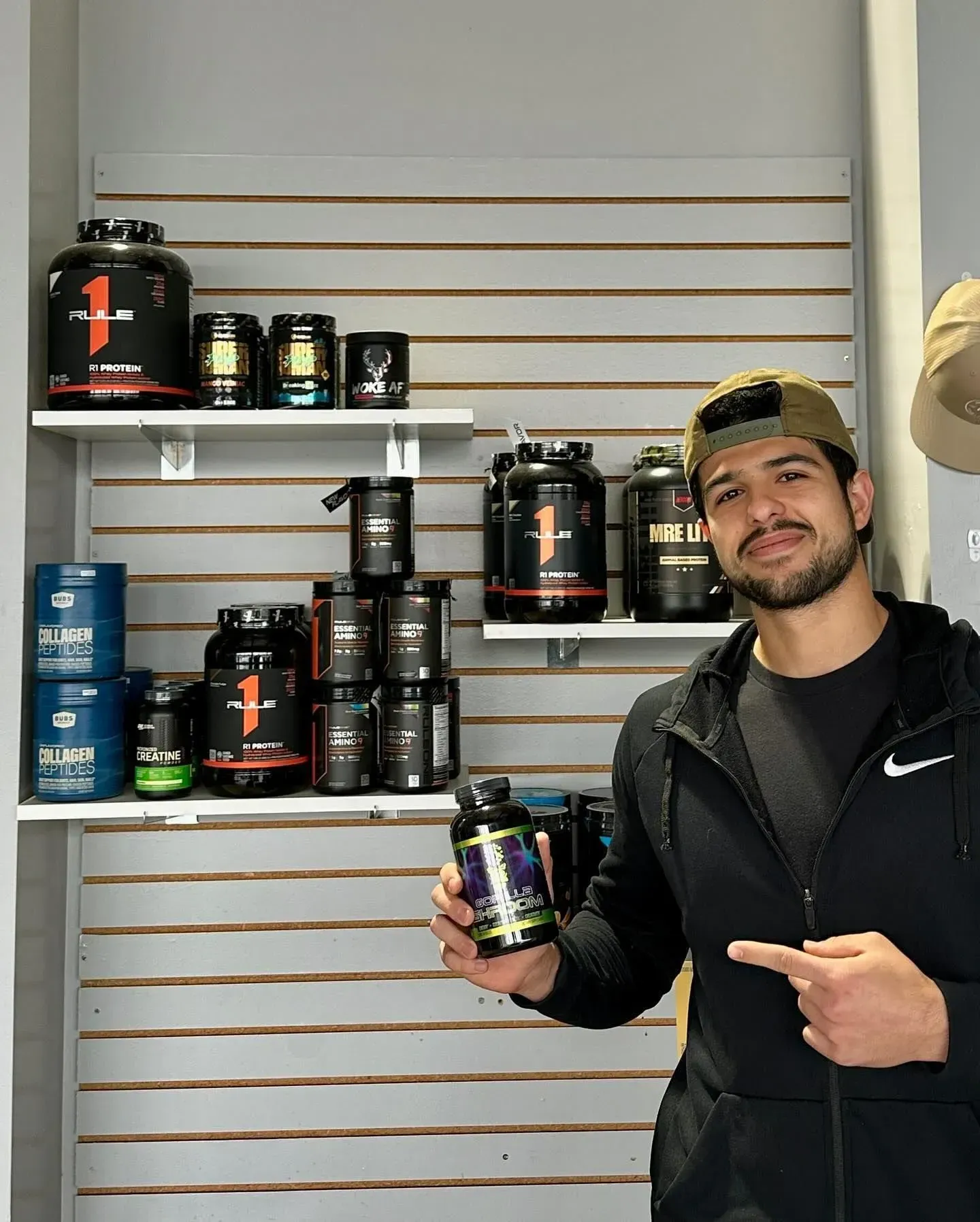 Man in a gym holding a supplement, pointing at it. Shelves behind him hold various supplement containers.