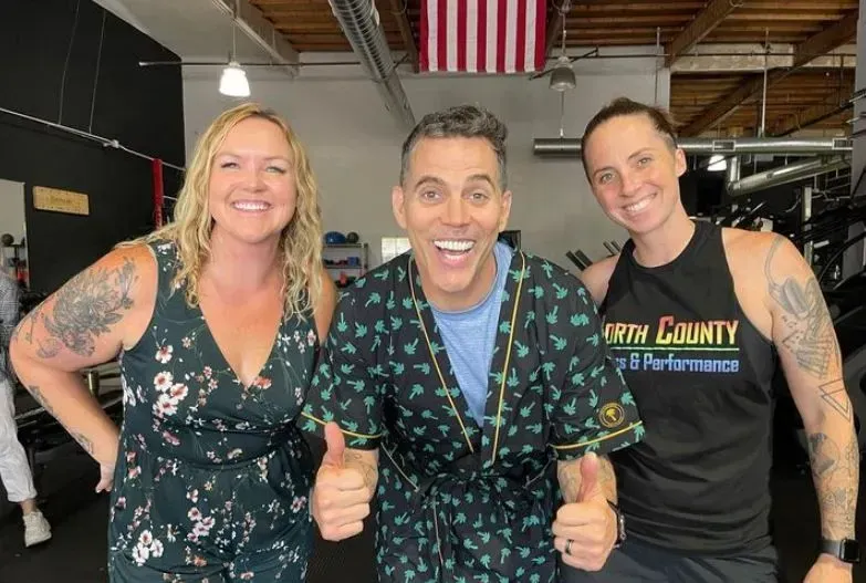Three people smiling in a gym. Man in robe gives thumbs up; other two have arms around each other. American flag.