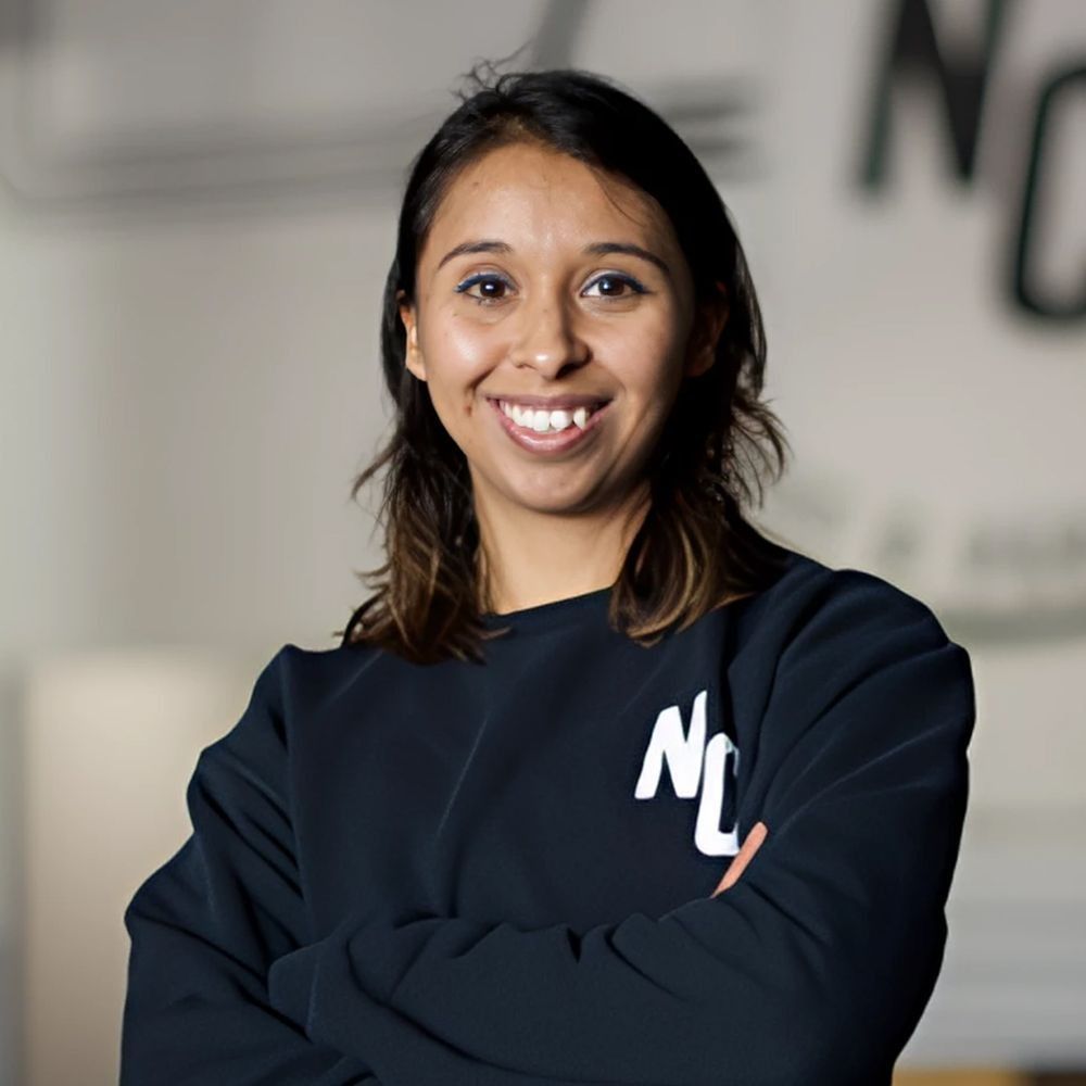 Woman with brown hair smiles, arms crossed, wearing a black sweatshirt with 