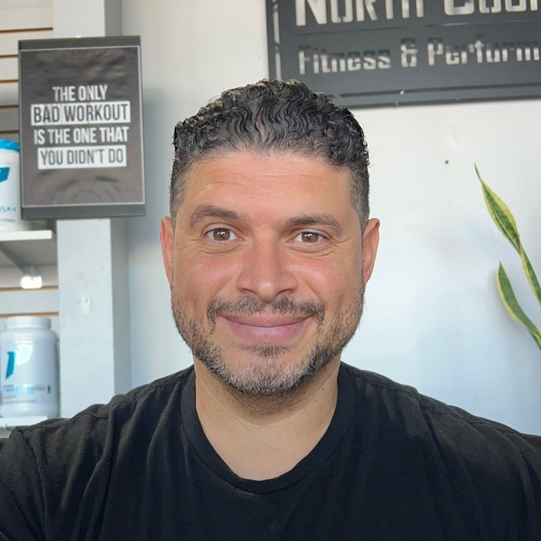 Man smiling, in a gym. Black t-shirt, short dark hair, and a trimmed beard. Sign: 