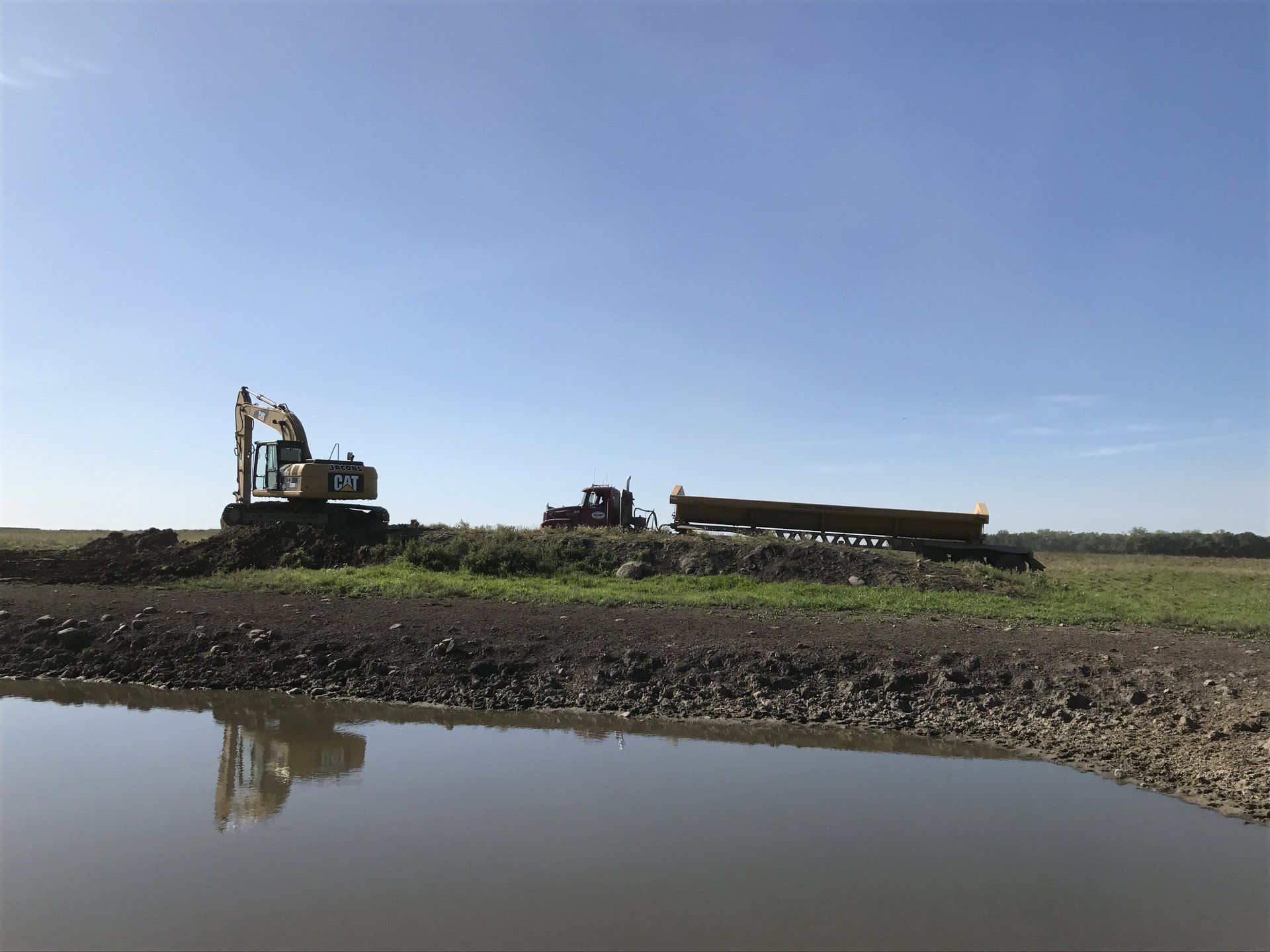 An excavator and a truck are working in a field next to a body of water.