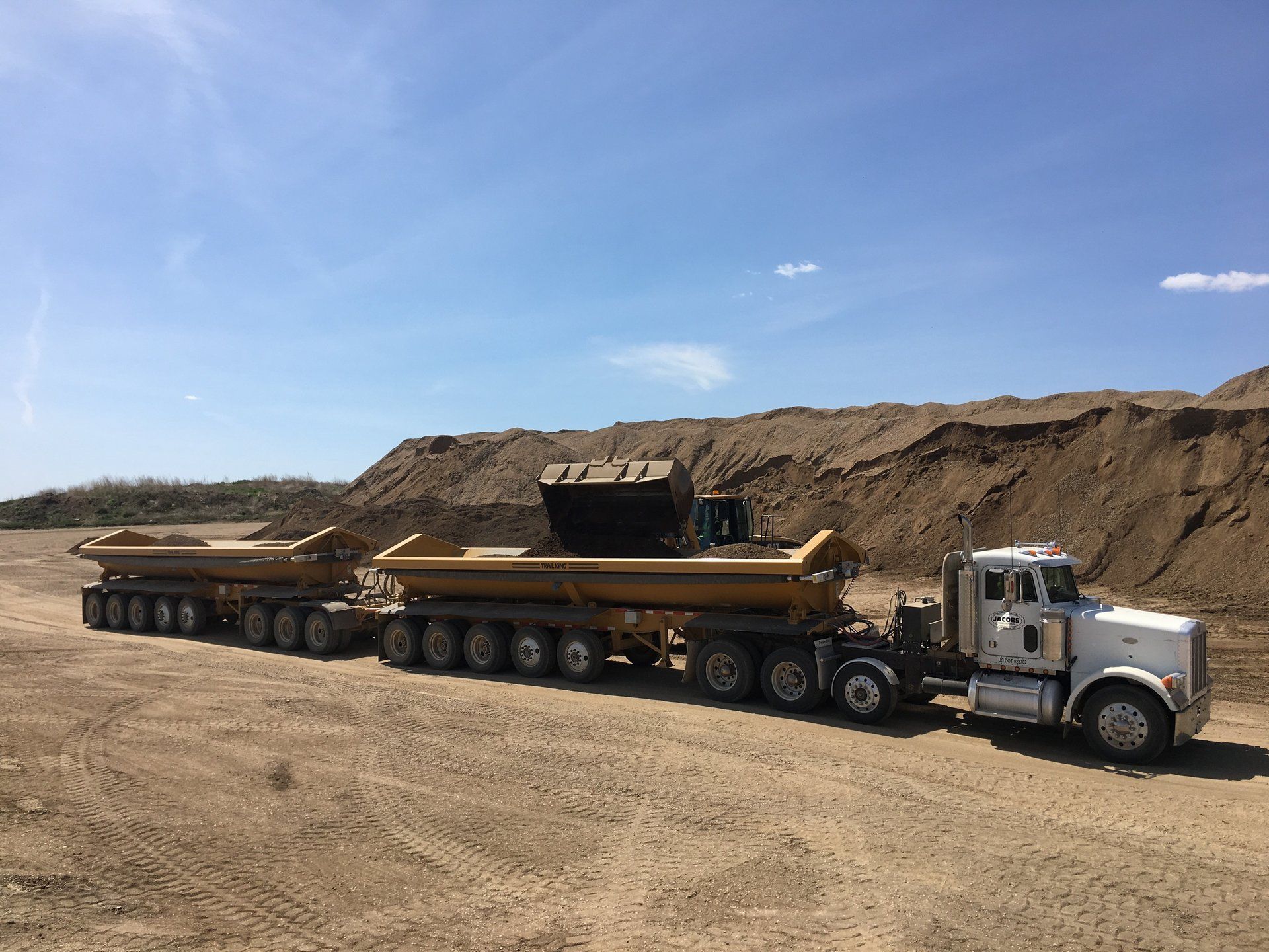 A semi truck is driving down a dirt road next to a pile of dirt.