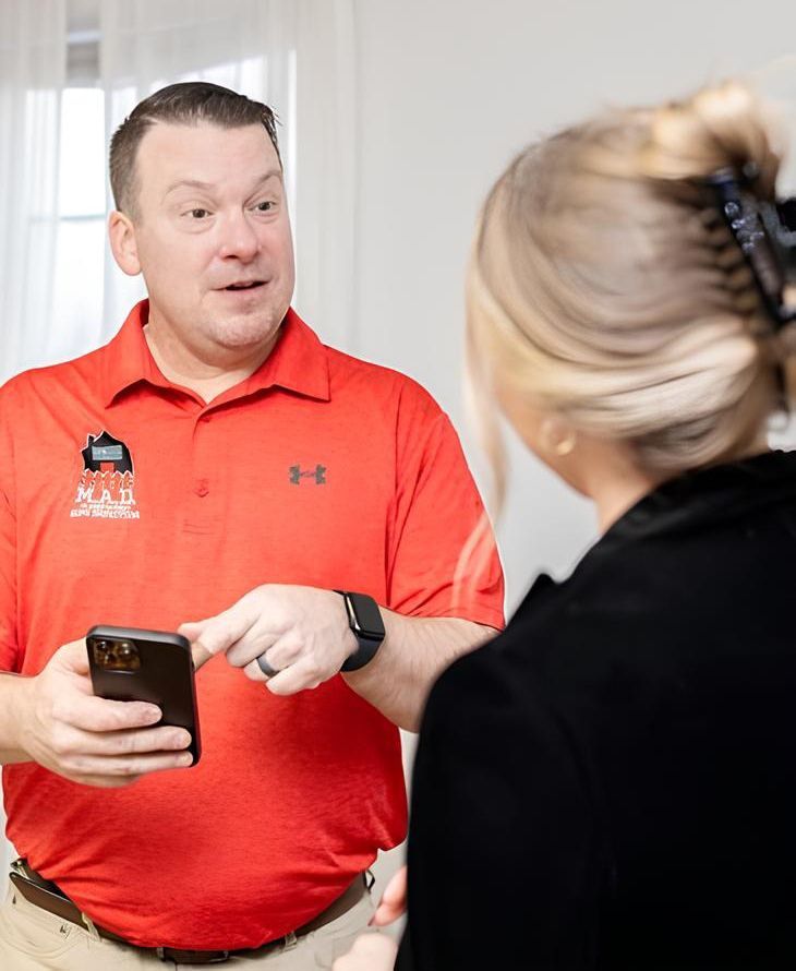 Man in red shirt points at phone, speaking to woman with blonde hair indoors.