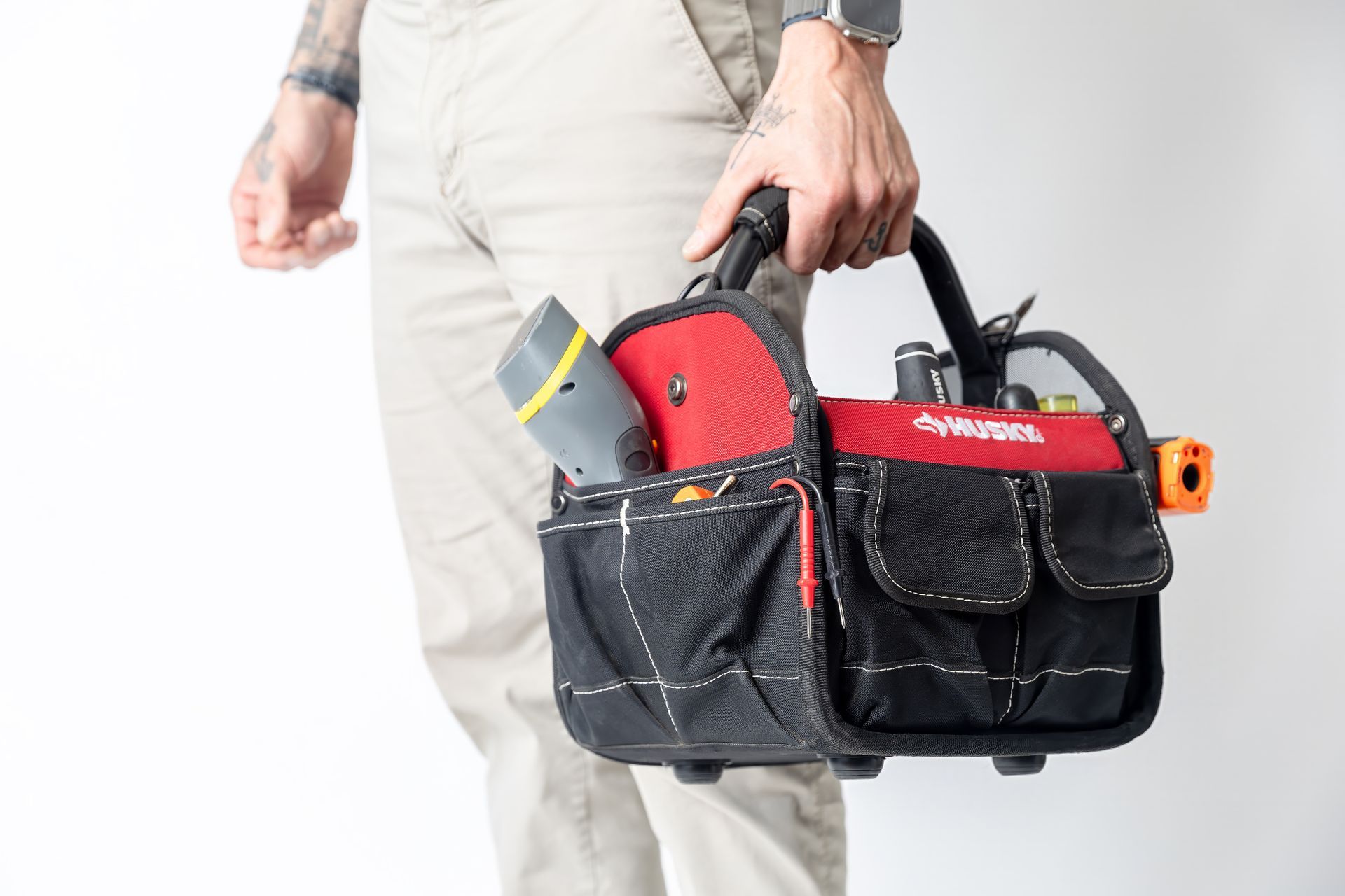 Person holding a red and black tool bag with tools in it. Beige pants and a white background.
