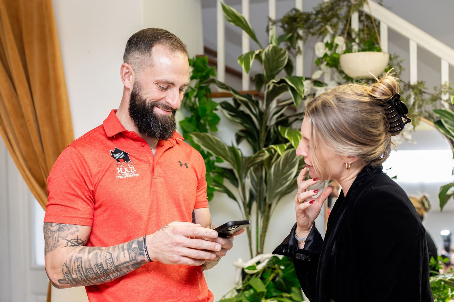 Man in orange shirt shows phone to woman indoors, plants in background.