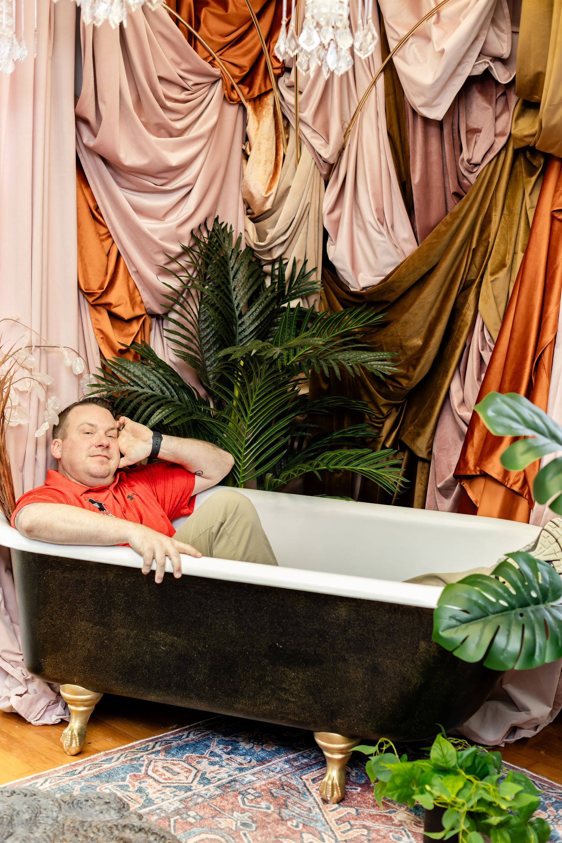Man in red shirt relaxed in a claw-foot tub, surrounded by draped fabrics and greenery.
