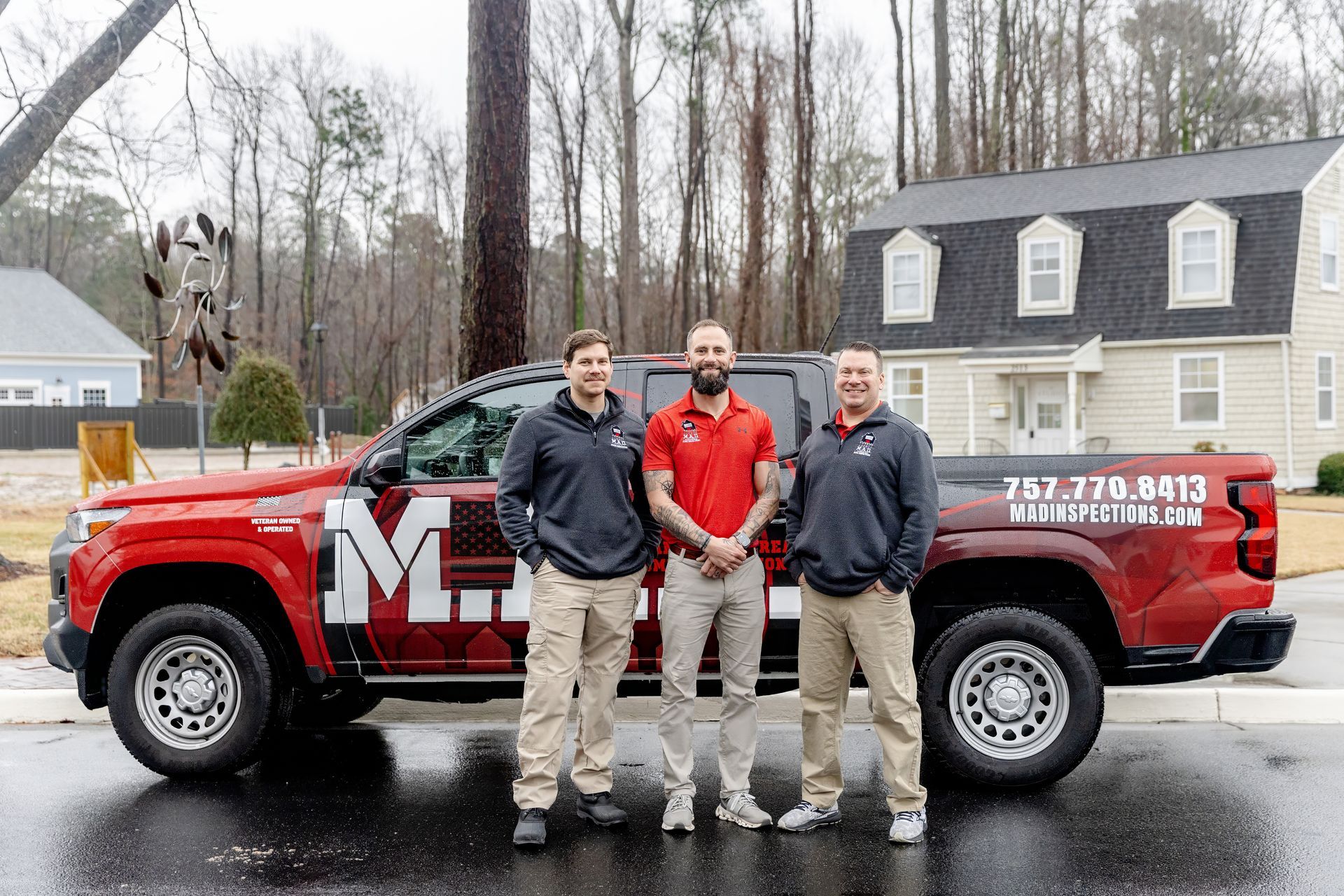 Three men stand next to a red truck with a logo. Cloudy day, residential setting.