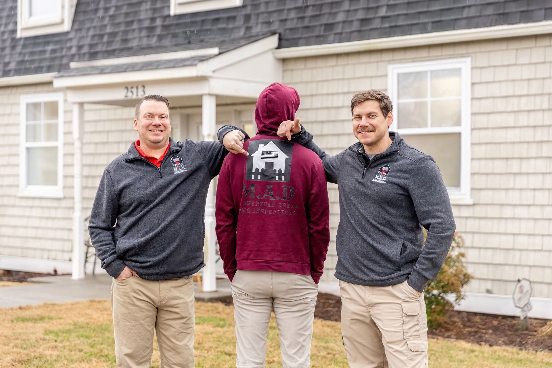 Three men in front of a house, two with arms on the third, who wears a hoodie. Gray, red, tan, and maroon clothing.
