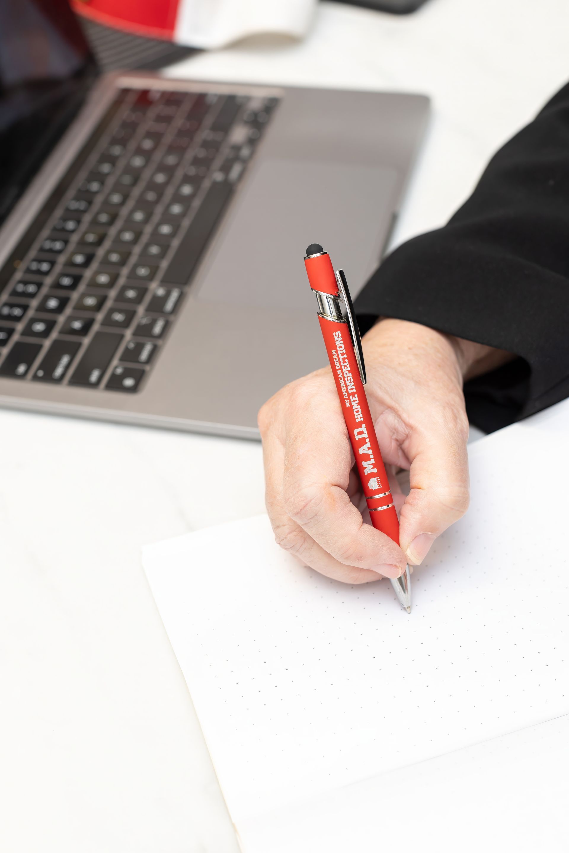 Person writing on paper with a red pen, next to a laptop on a white desk.