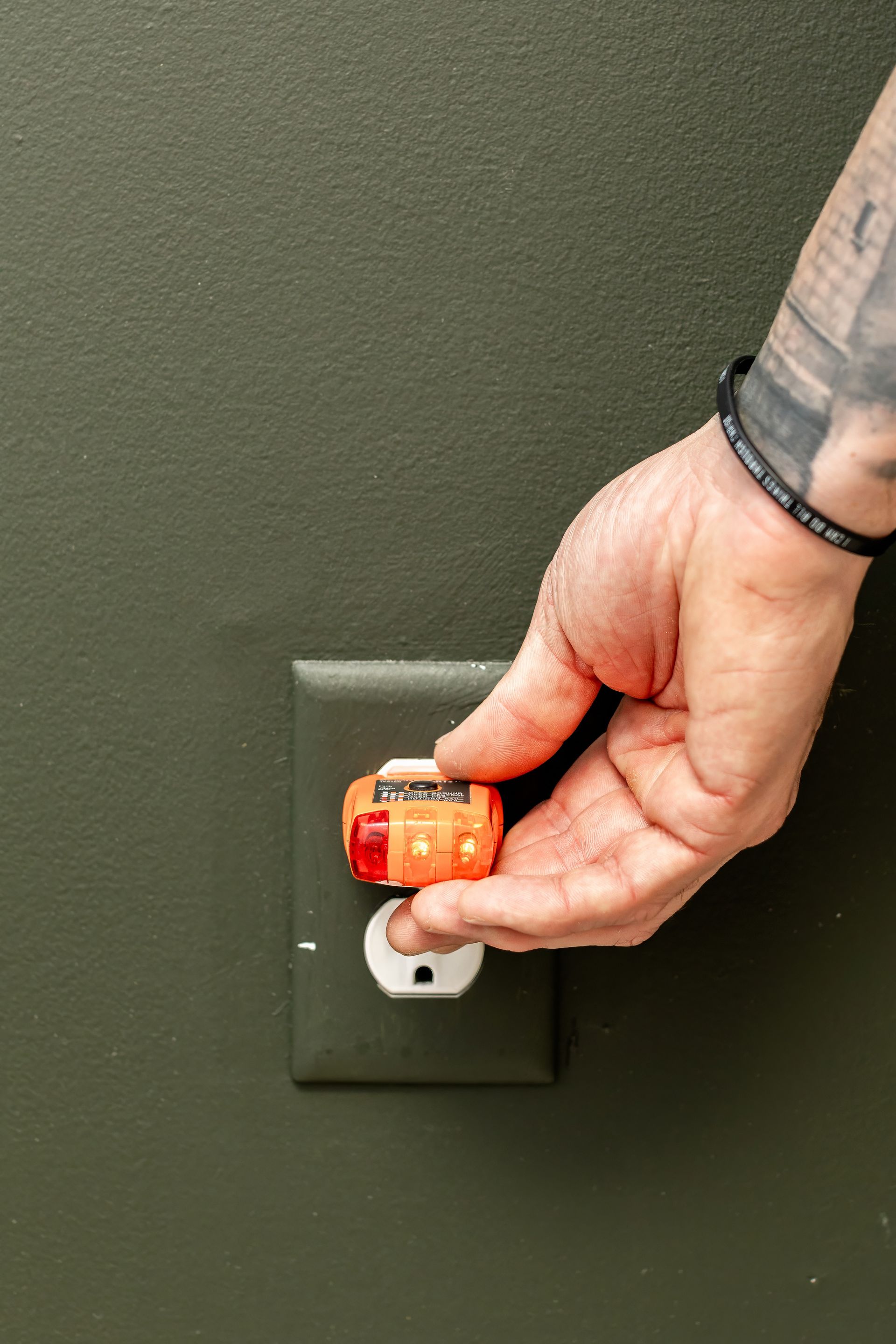 Hand inserting an orange timer into a green electrical outlet on a dark green wall.