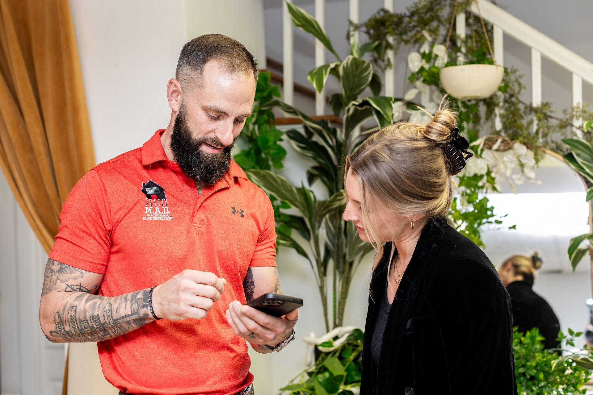 Man in orange shirt shows woman phone. They are surrounded by plants and near stairs.