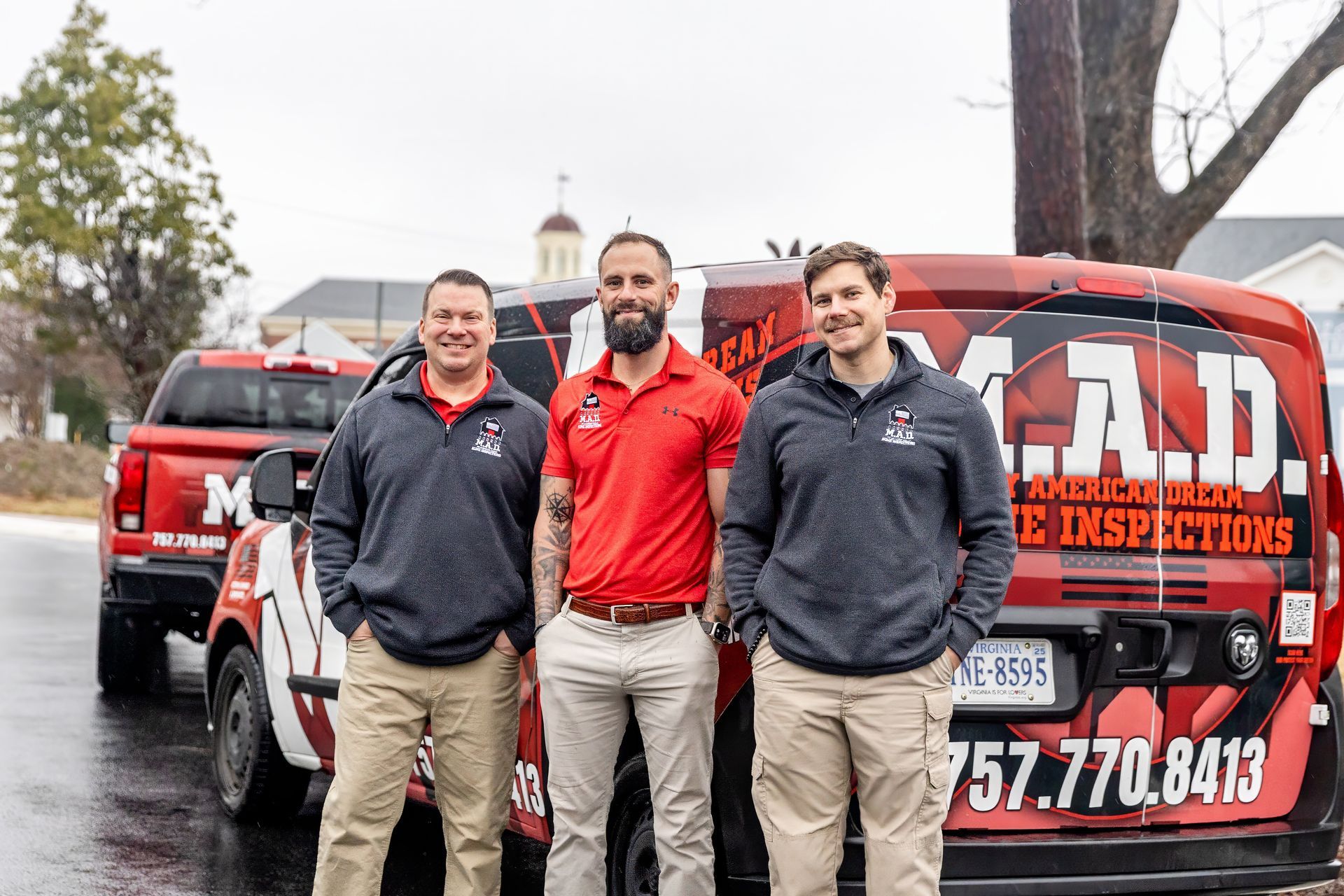Three men stand near red and white work vehicles. One wears a red shirt. Others wear grey sweatshirts and khaki pants.