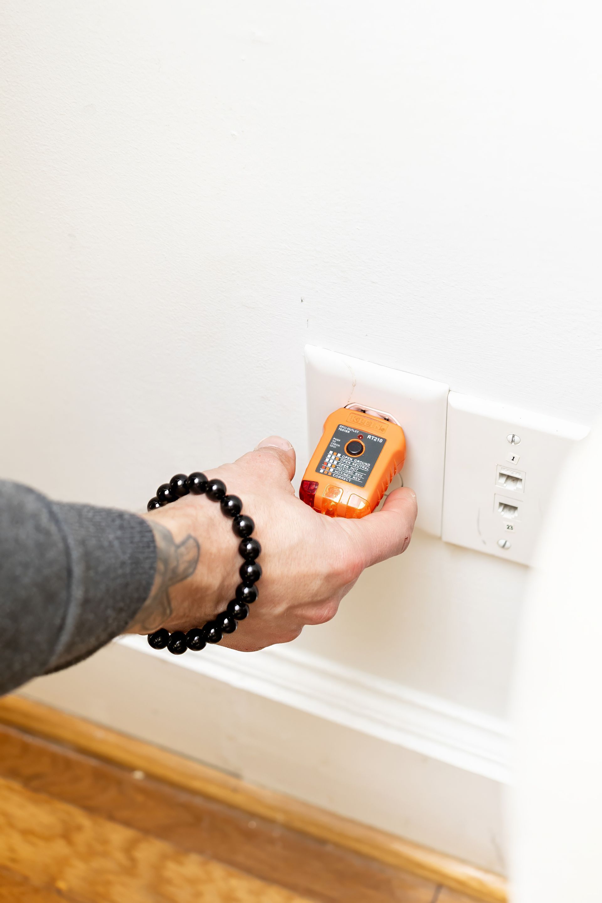 Hand testing an electrical outlet with an orange circuit tester. White wall, wooden baseboard.