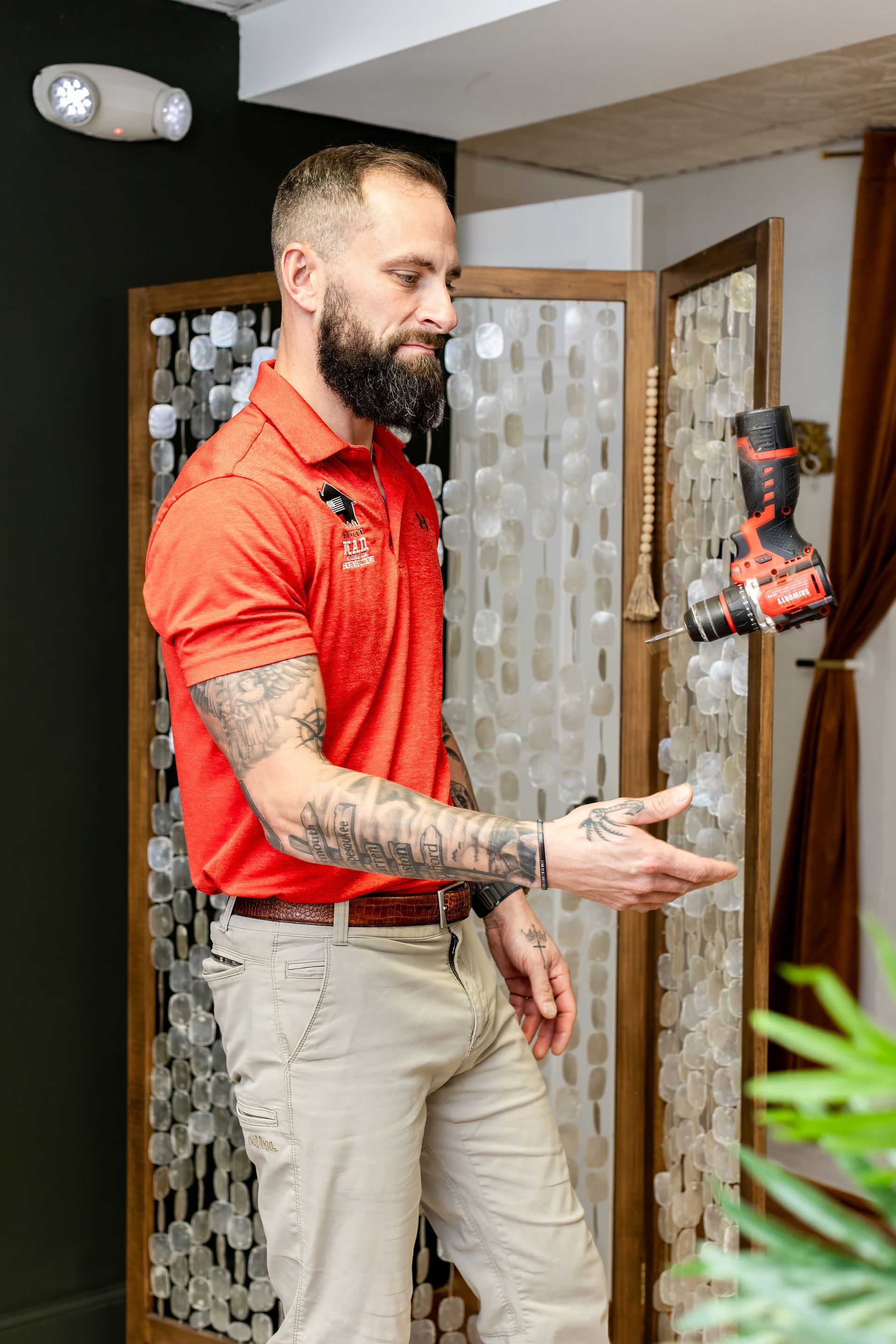 Man in red shirt points toward a suspended drill near a beaded screen in an office.