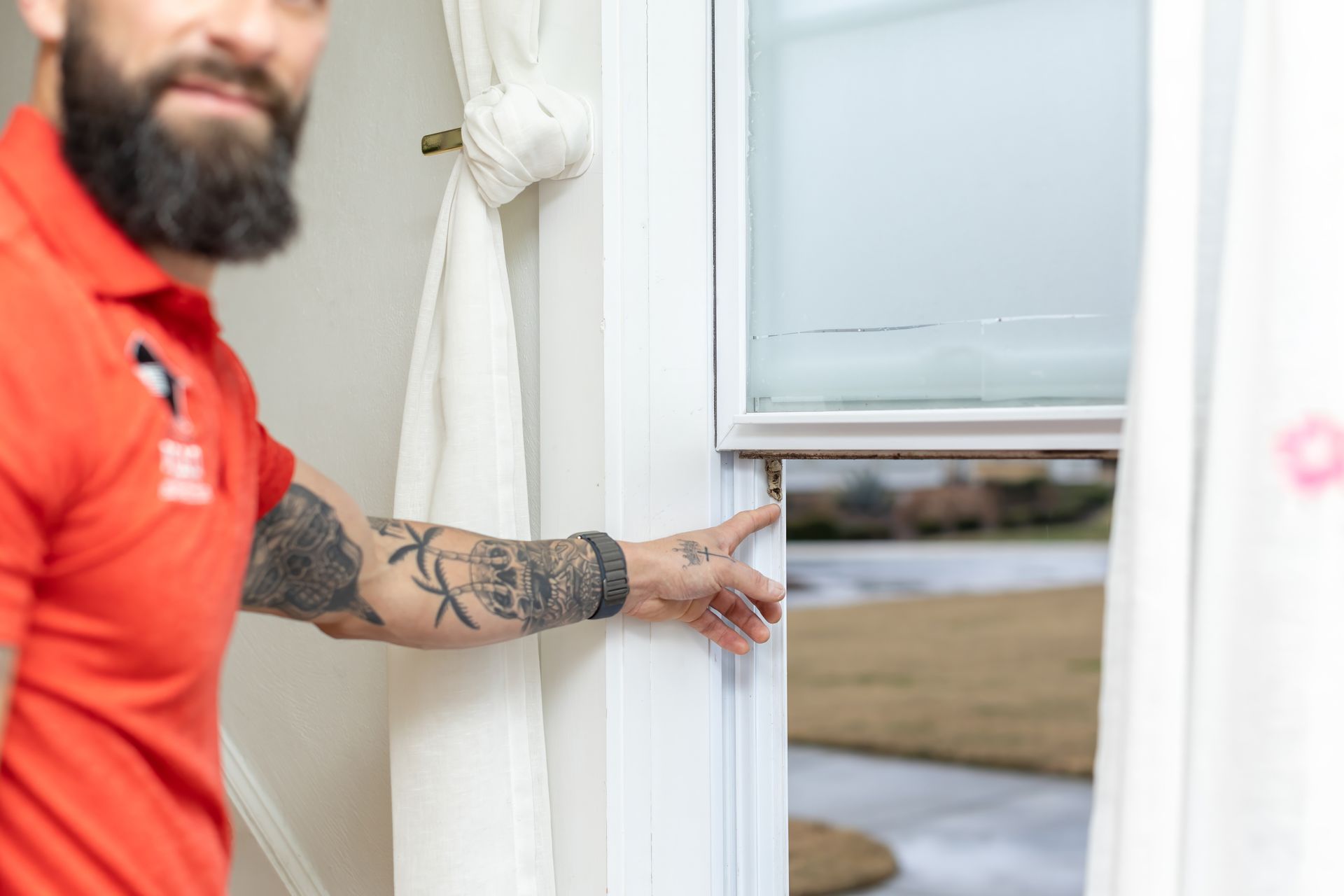 Man in red shirt touching a window frame. He has a beard and arm tattoos; a white curtain is to the left.