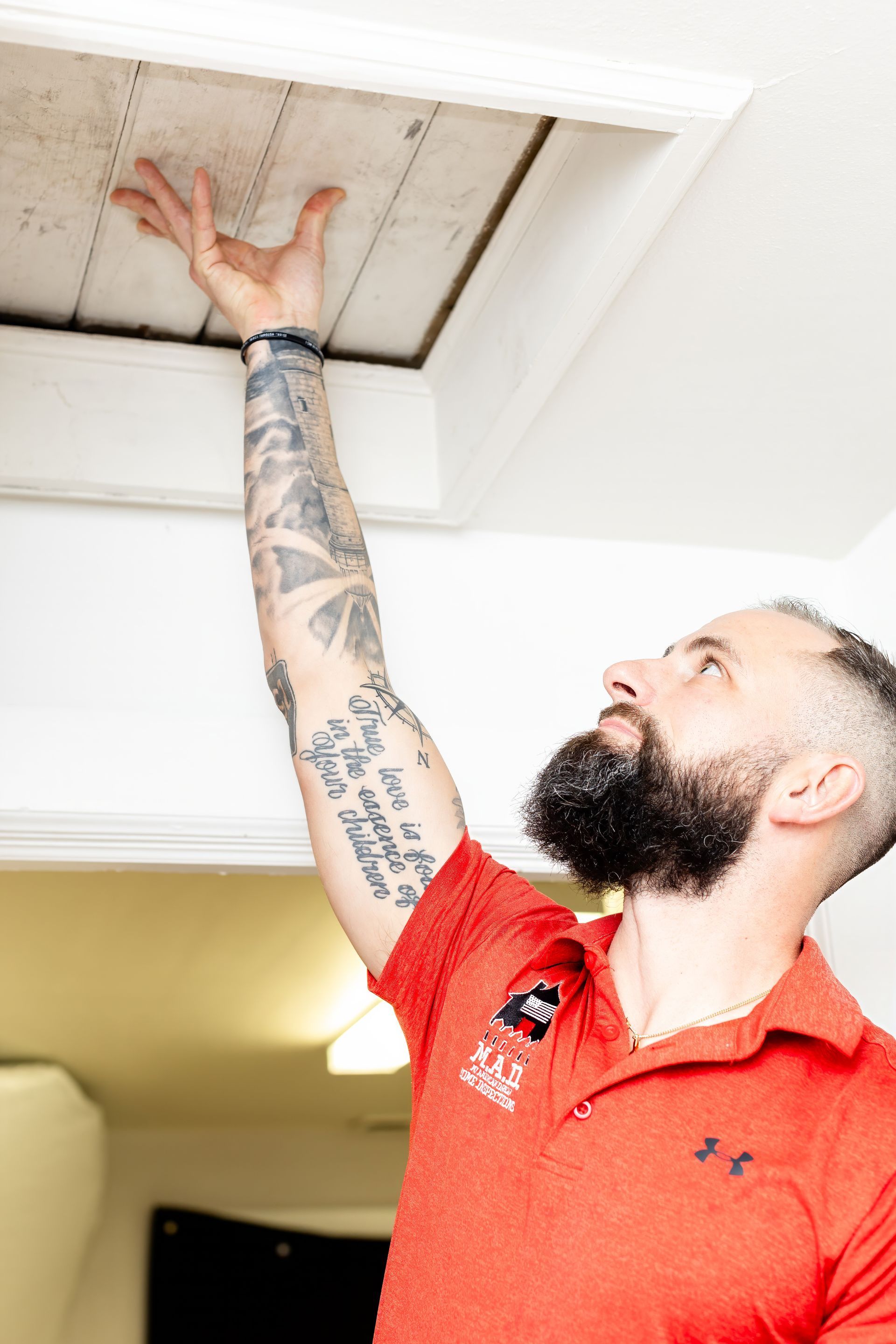 Man reaching up to open attic access panel. He has tattoos and a beard, wearing a red shirt.
