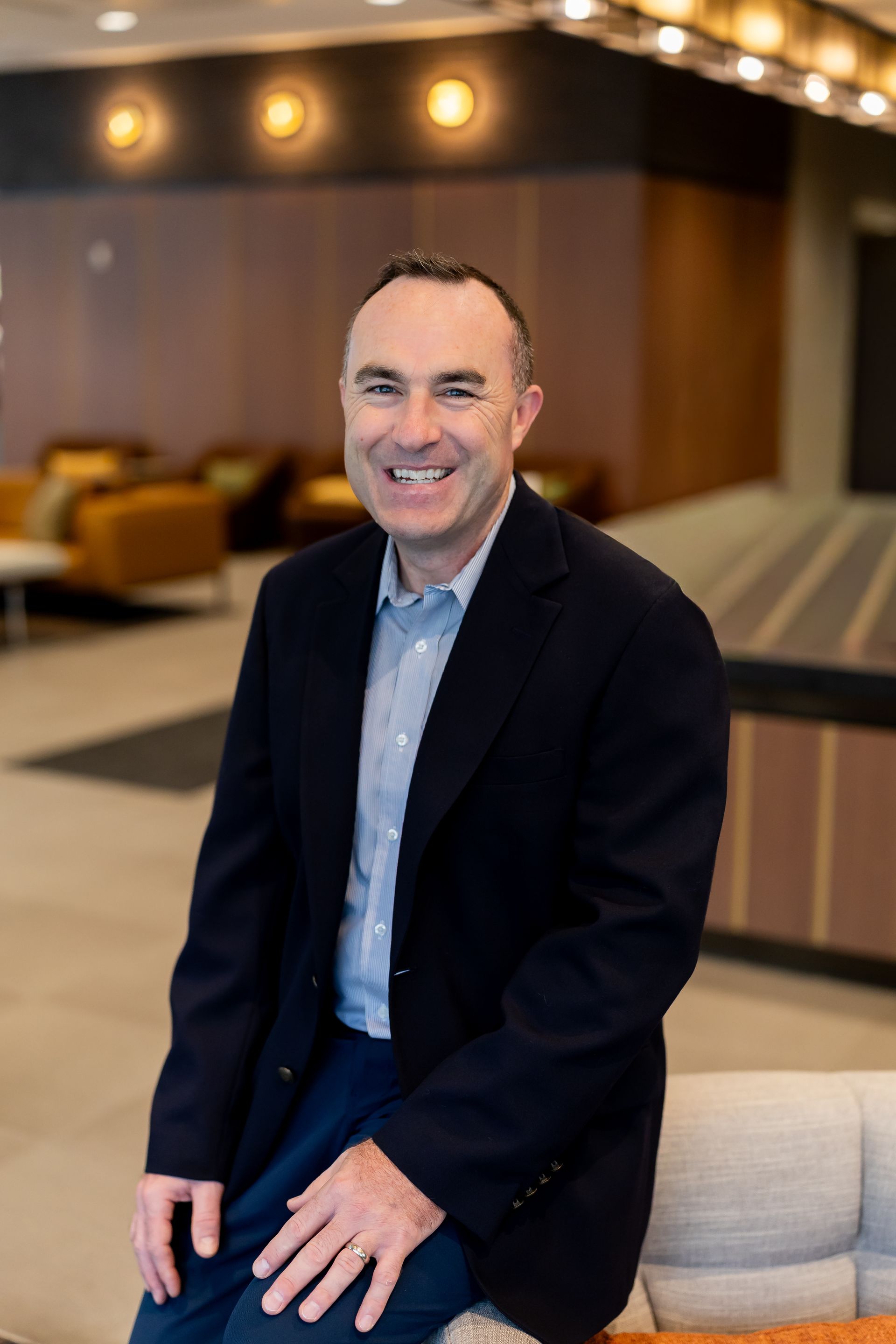 Smiling man in a dark blazer standing in a modern lobby with warm lighting