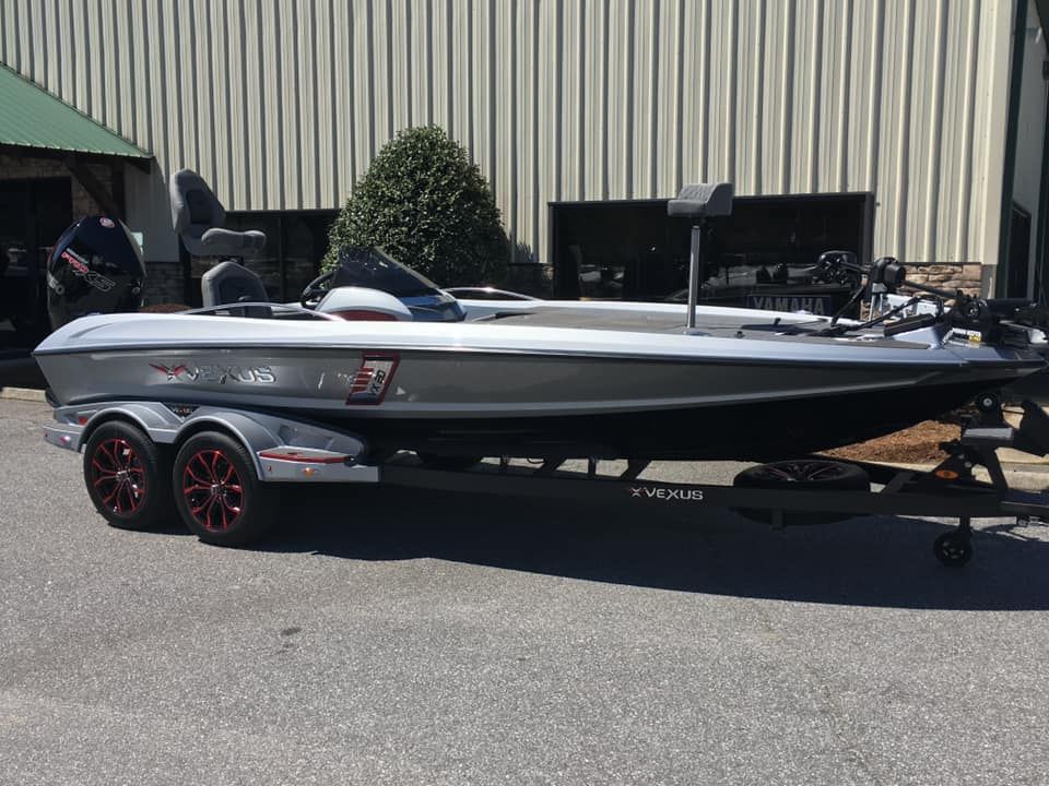 Silver bass boat on a trailer, parked on pavement in front of a building on a sunny day.
