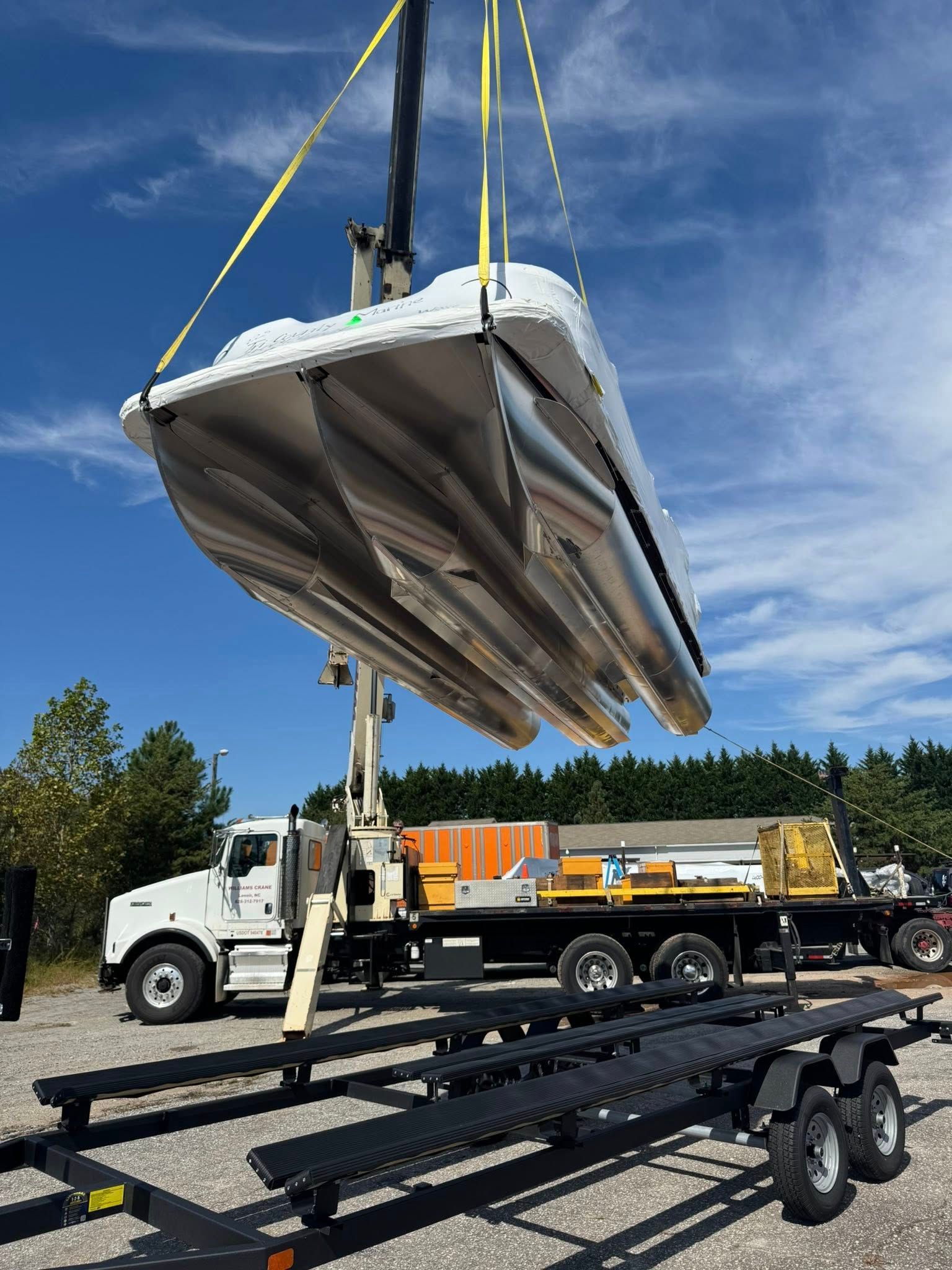 A pontoon boat is lifted by a crane onto a flatbed truck on a sunny day.