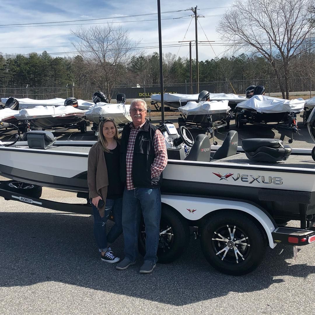 A man and woman standing by a new white and black Vexus boat; parked outdoors.