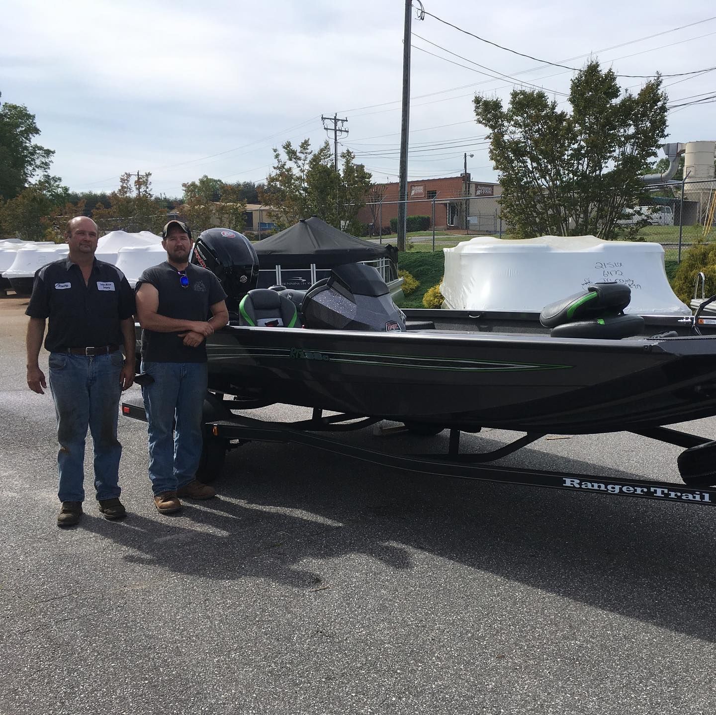 Two men stand near a dark boat on a trailer, in a parking lot. Cloudy sky, other boats covered.