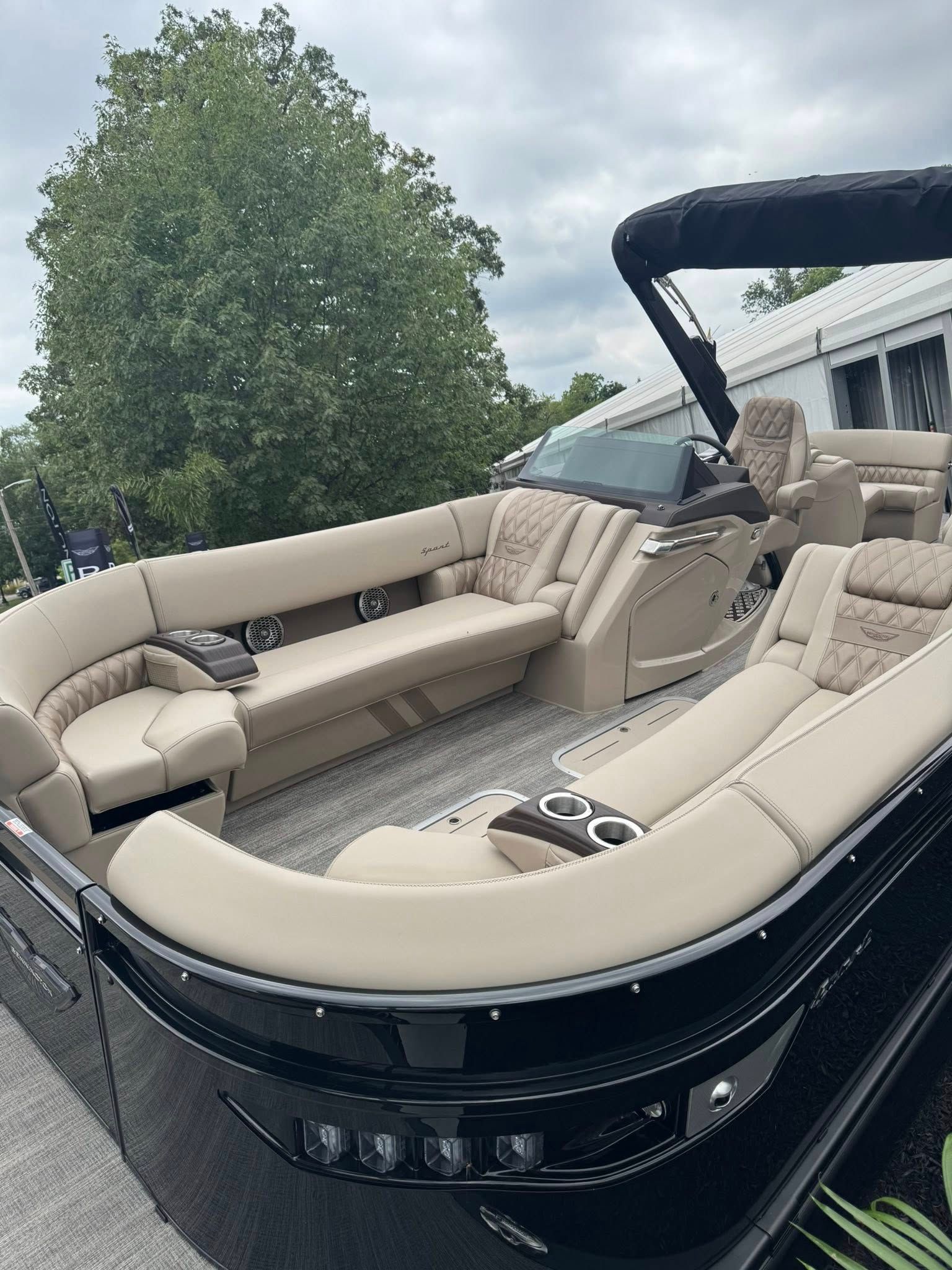 Black and beige pontoon boat on land with plush seating, cup holders, and a black canopy.