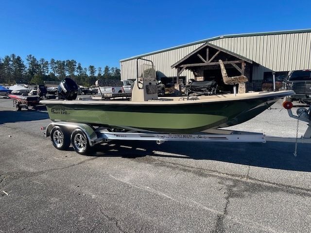 Green and tan fishing boat on a trailer, parked on a paved lot. A building is in the background.