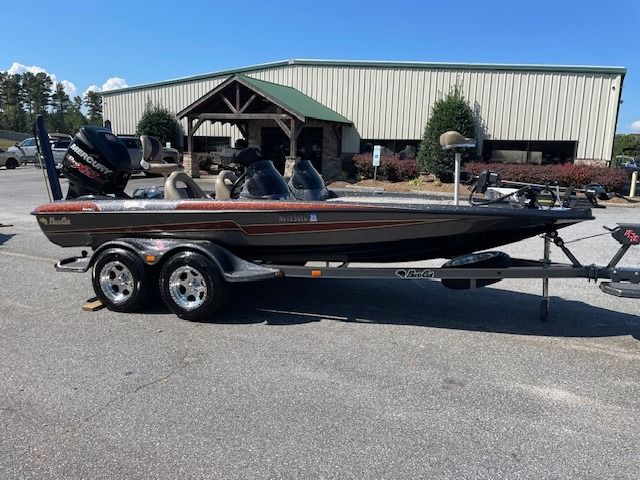 Brown bass boat on a trailer parked in a paved lot in front of a building with a green roof.
