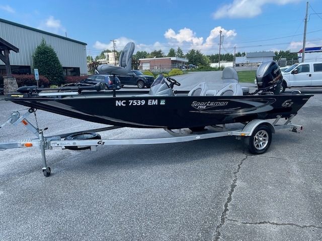 Black fishing boat on a trailer, parked on pavement. Boat has a motor and two seats.