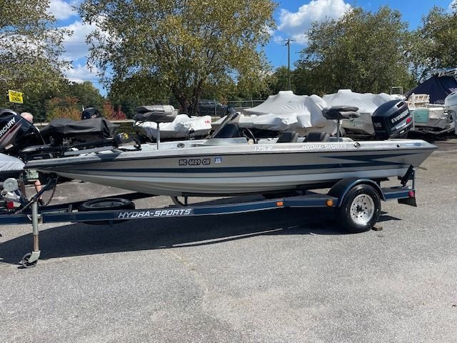 Silver and blue Hydra-Sports bass boat on trailer in parking lot, trees in background.