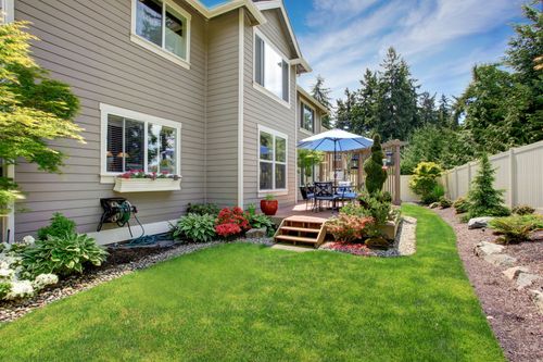 House with landscaped backyard, green lawn, flower beds, and patio with umbrella and steps
