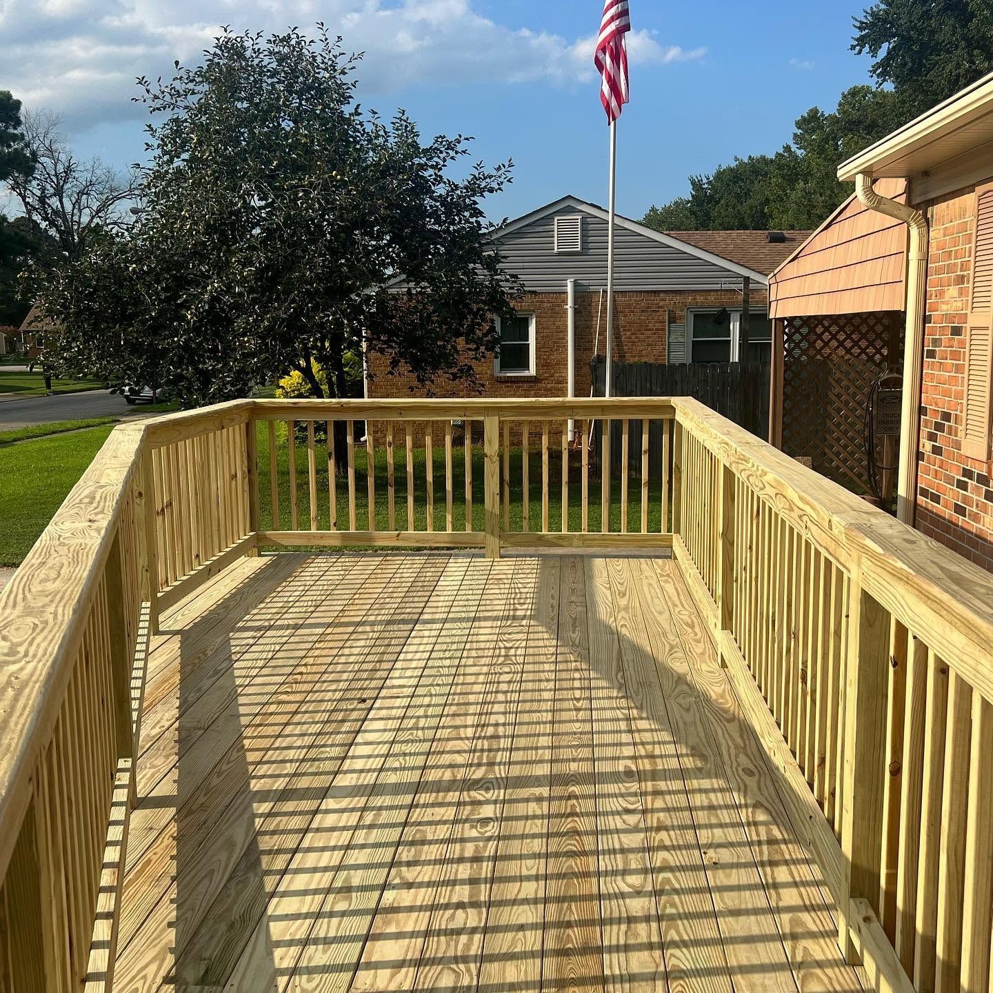 Wooden backyard deck with railing, overlooking a brick house, lawn, and flagpole on a sunny day