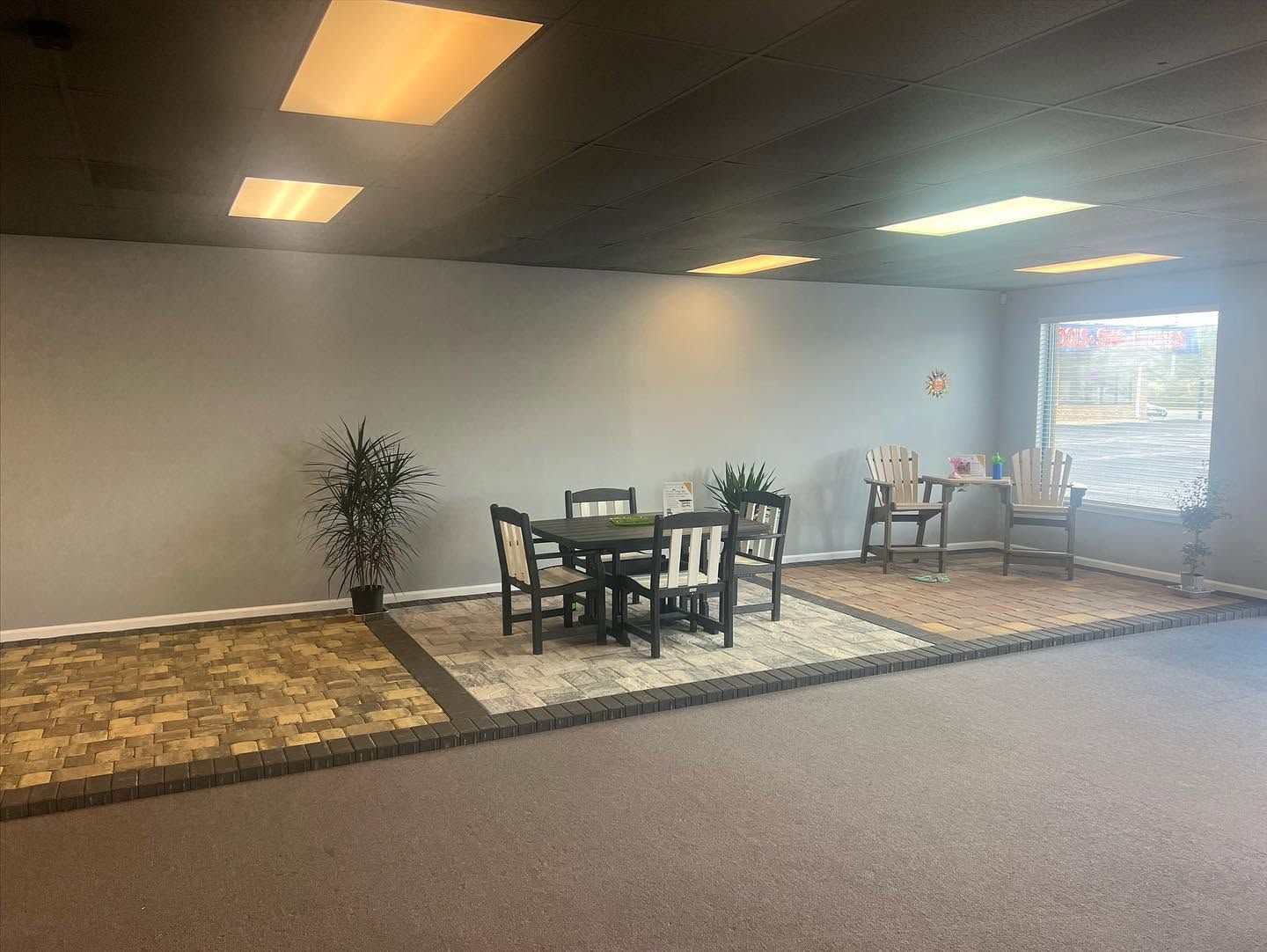 Empty office lounge with tables, chairs, potted plants, and bright ceiling lights near a window.