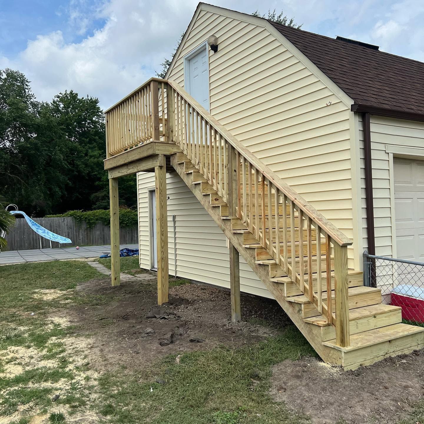 Wooden exterior staircase and deck attached to a beige house on posts in a backyard.