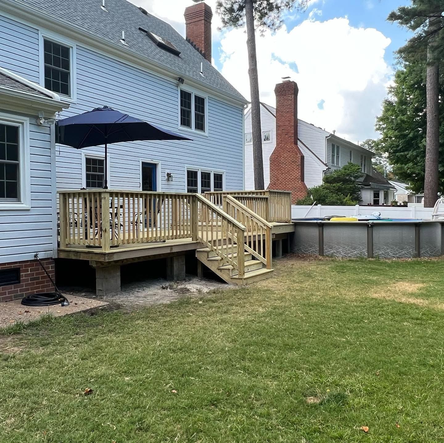 Backyard deck with stairs and a blue umbrella beside a white house, overlooking a grassy lawn