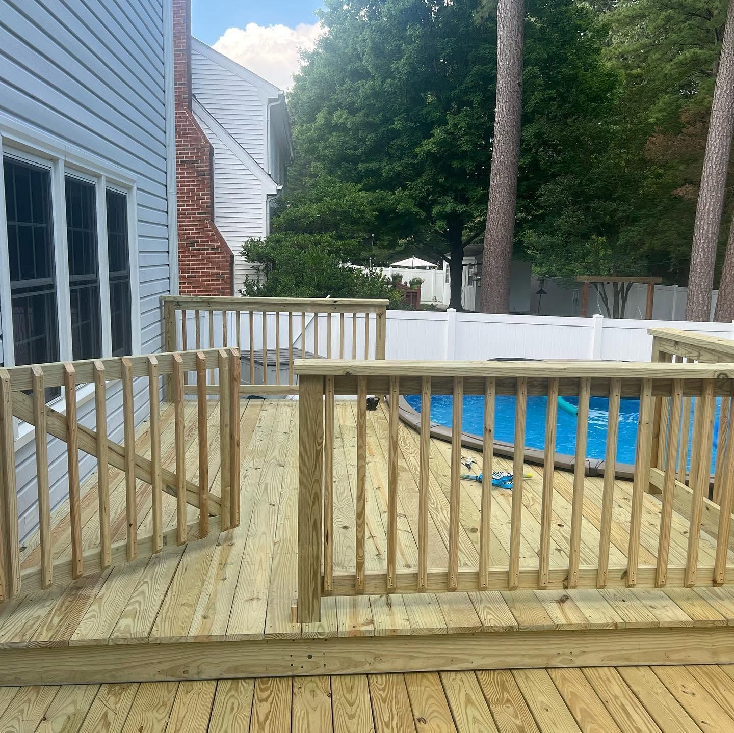 Wooden deck with railing overlooking an in-ground pool beside a house and backyard trees