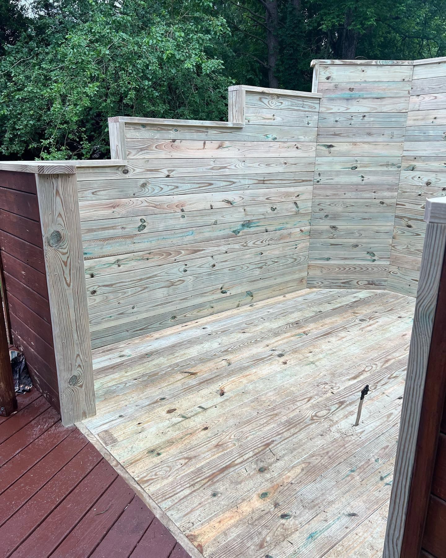 Wooden outdoor shower stall with stepped ledges on a red deck, backed by trees.