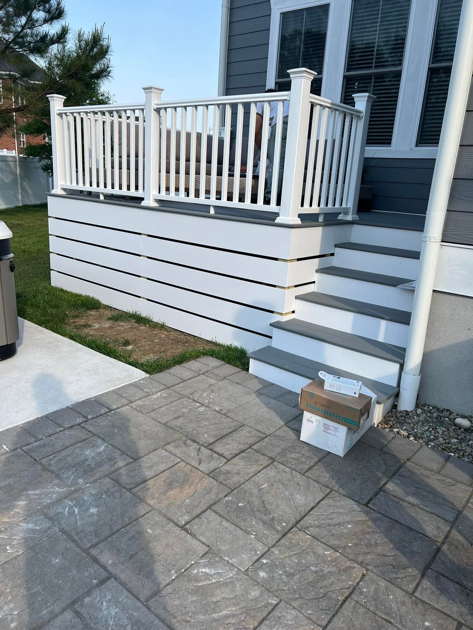White house porch with stairs, railing, and a cardboard box on the paved driveway.