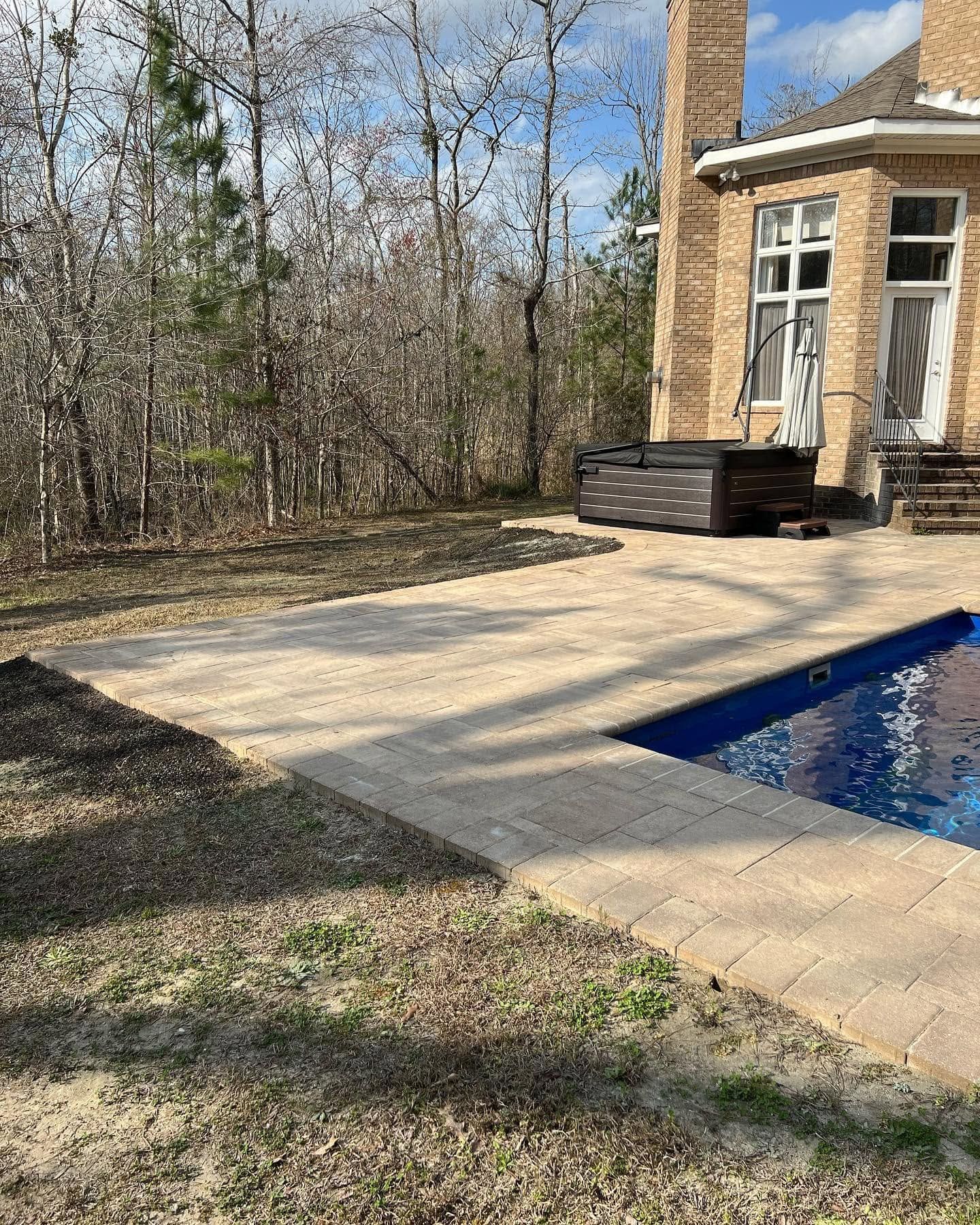Patio beside a brick house with a pool, stone paving, and a black hot tub near bare trees
