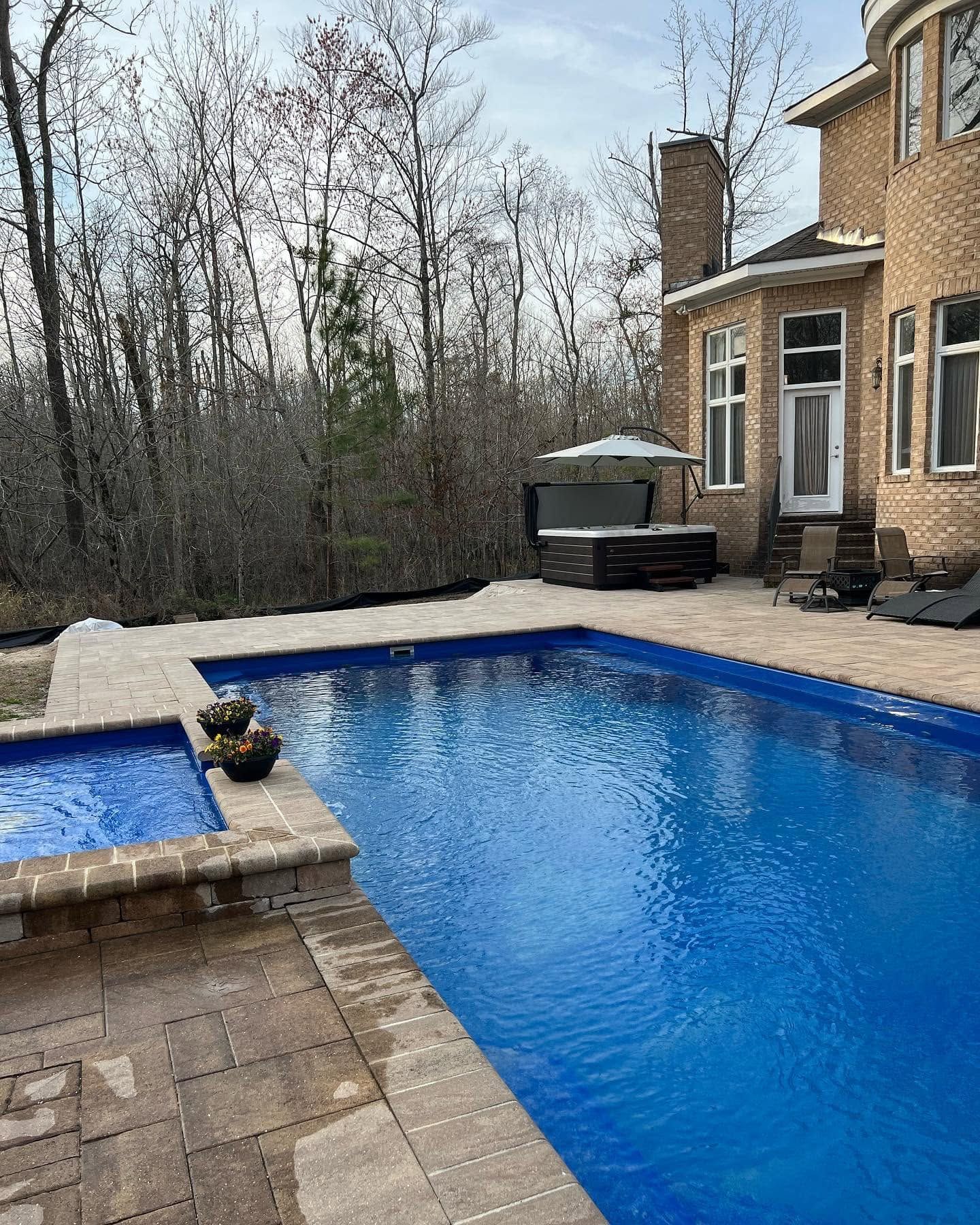 Backyard patio with blue pool beside a brick house and woods in the background