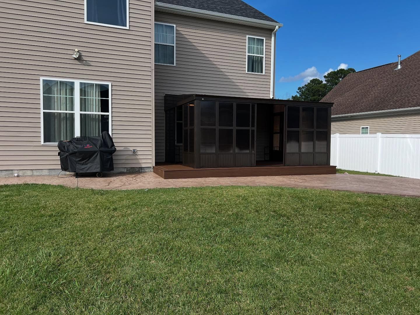Backyard patio beside a beige two-story house with a grassy lawn and white fence