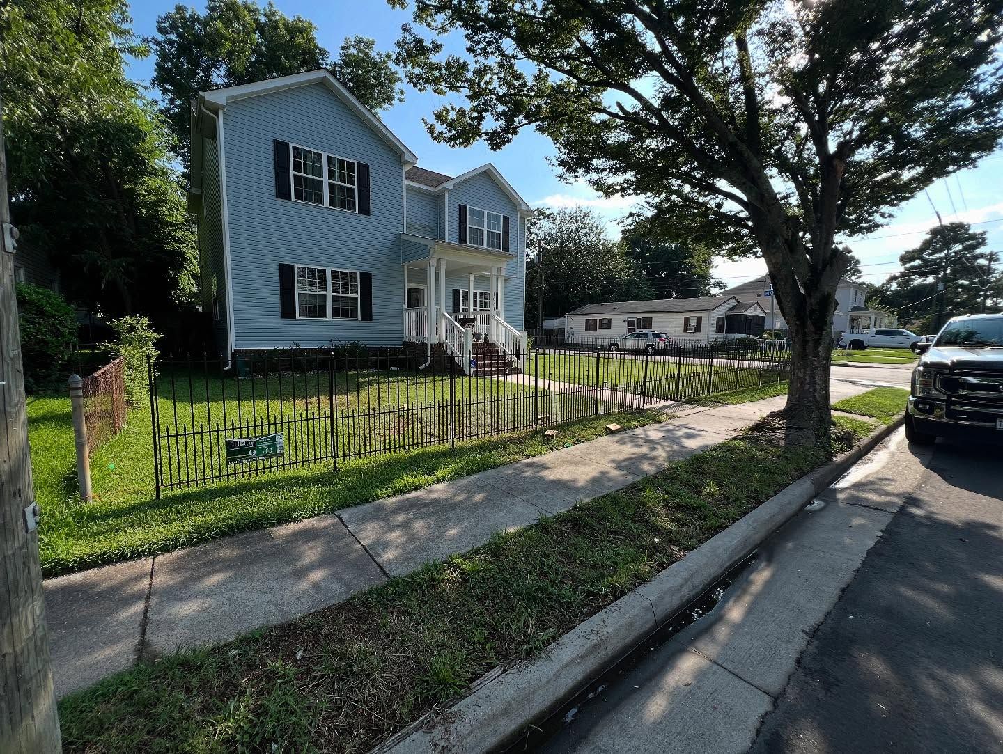 Blue house with a front porch beside a sidewalk, fence, and parked car on a sunny street.
