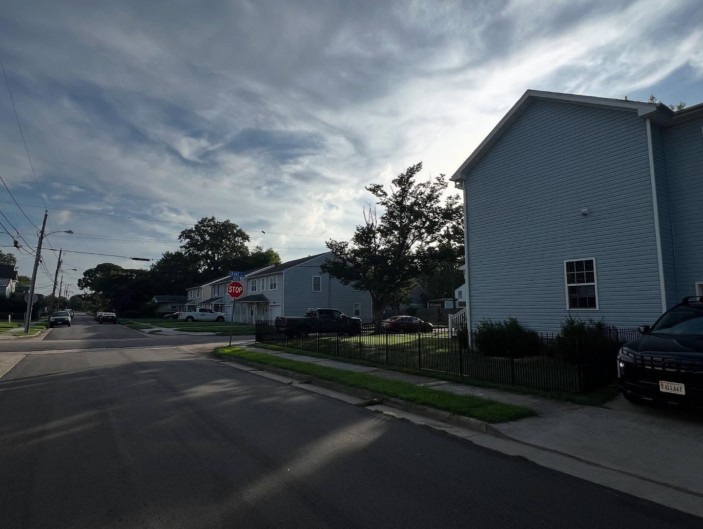 Quiet suburban street with white houses, trees, and a parked car under a cloudy sky