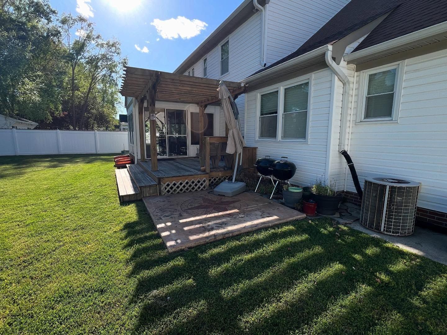 Backyard patio with a wooden pergola beside a white house, lit by low evening sunlight.