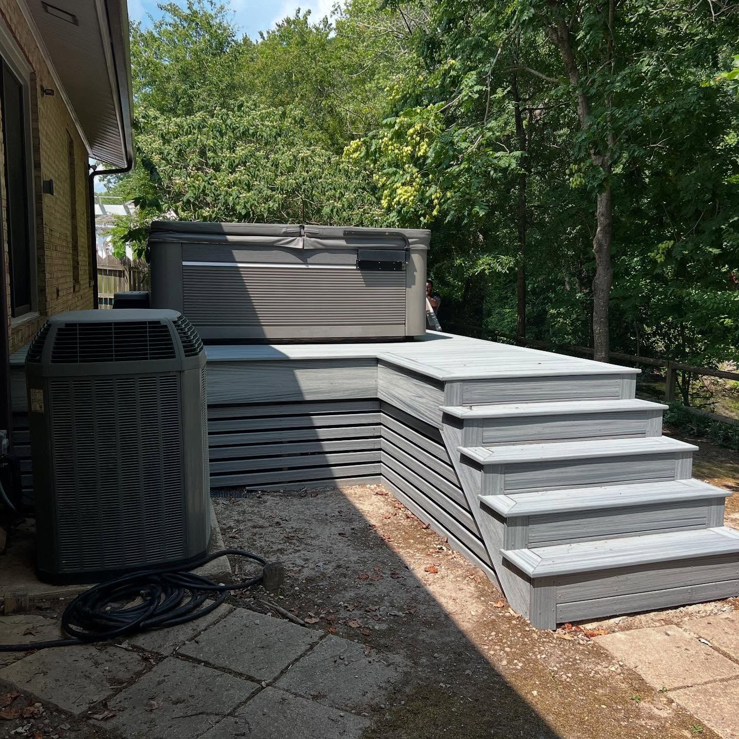 Backyard with gray multi-level deck steps beside a house and hot tub, surrounded by trees