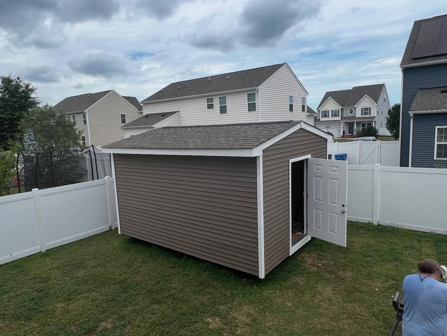 Backyard with a beige storage shed, white fence, and person kneeling near the corner